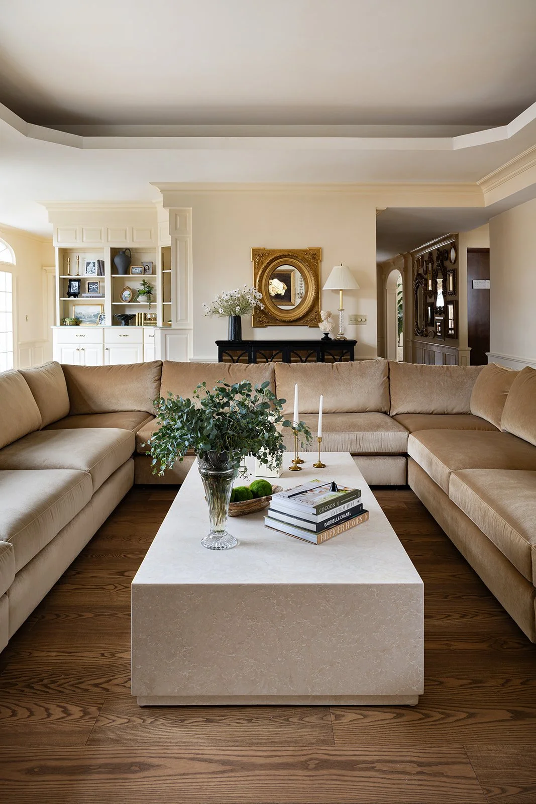 Living room with beige sectional sofa, white coffee table with books, green plant, candles, in front of a cream-colored wall with a gold-framed mirror and decorative items on a dark console table.