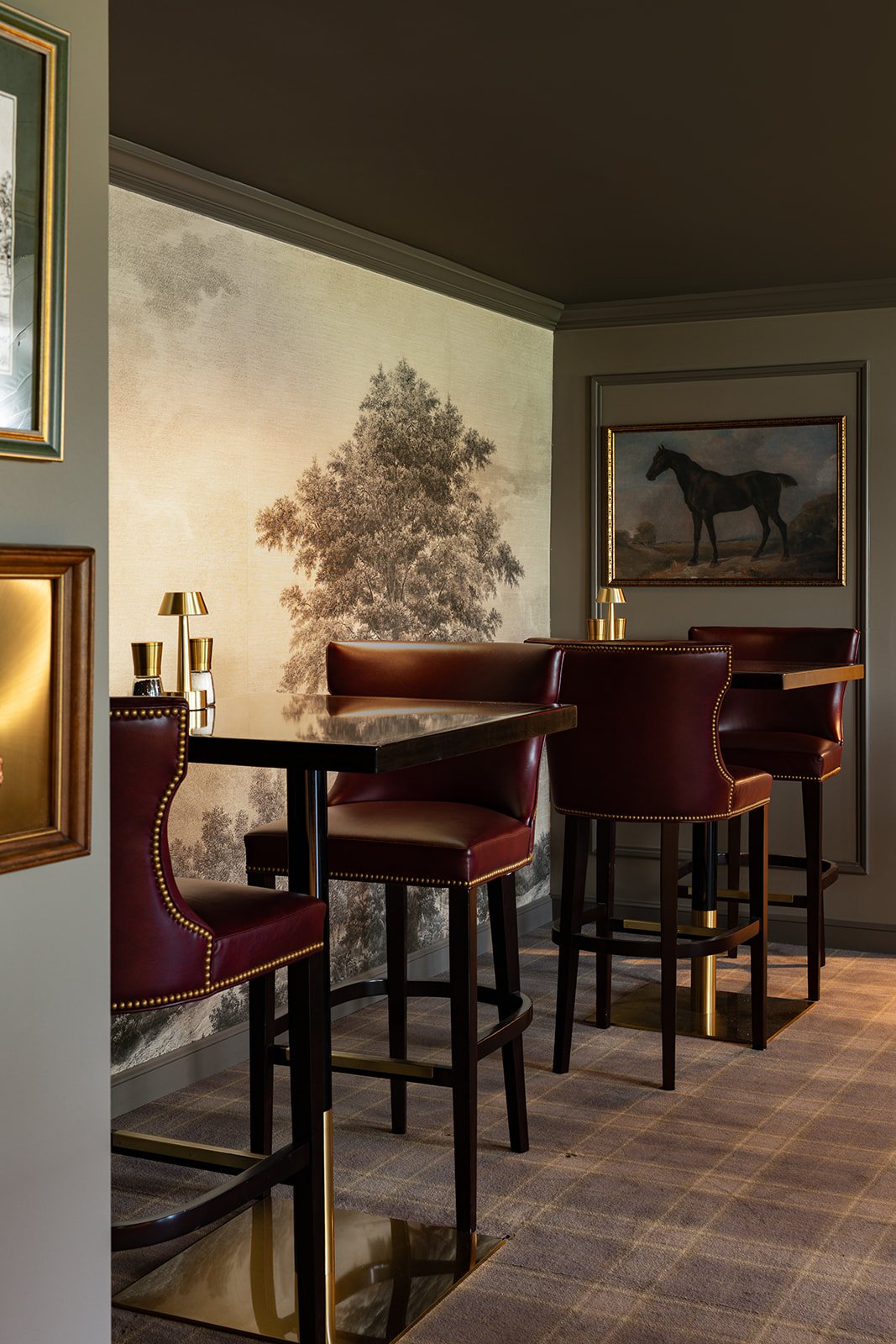 A cozy corner of a restaurant or lounge with high-backed burgundy leather chairs, a dark wooden table, gold lamp fixtures, framed horse paintings, and a large tree mural on the wall.
