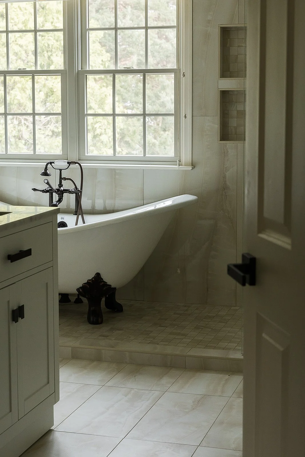 A bathroom with a clawfoot bathtub, large window, beige tile walls, and a built-in shower niche.