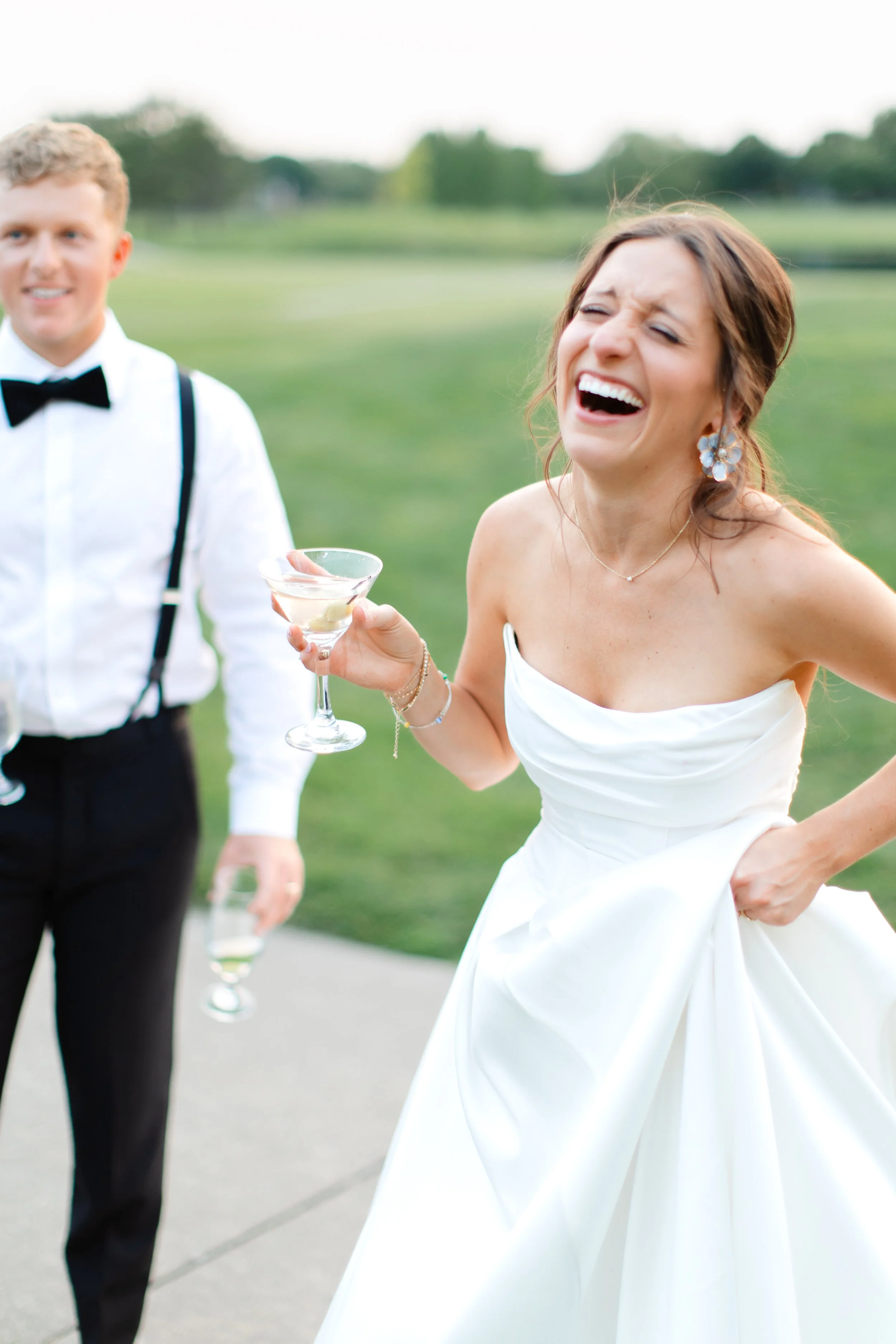 A woman in a wedding dress laughing and holding a glass of champagne, with a man in a tuxedo with suspenders and a bow tie standing nearby, in an outdoor setting with a grassy field and trees in the background.