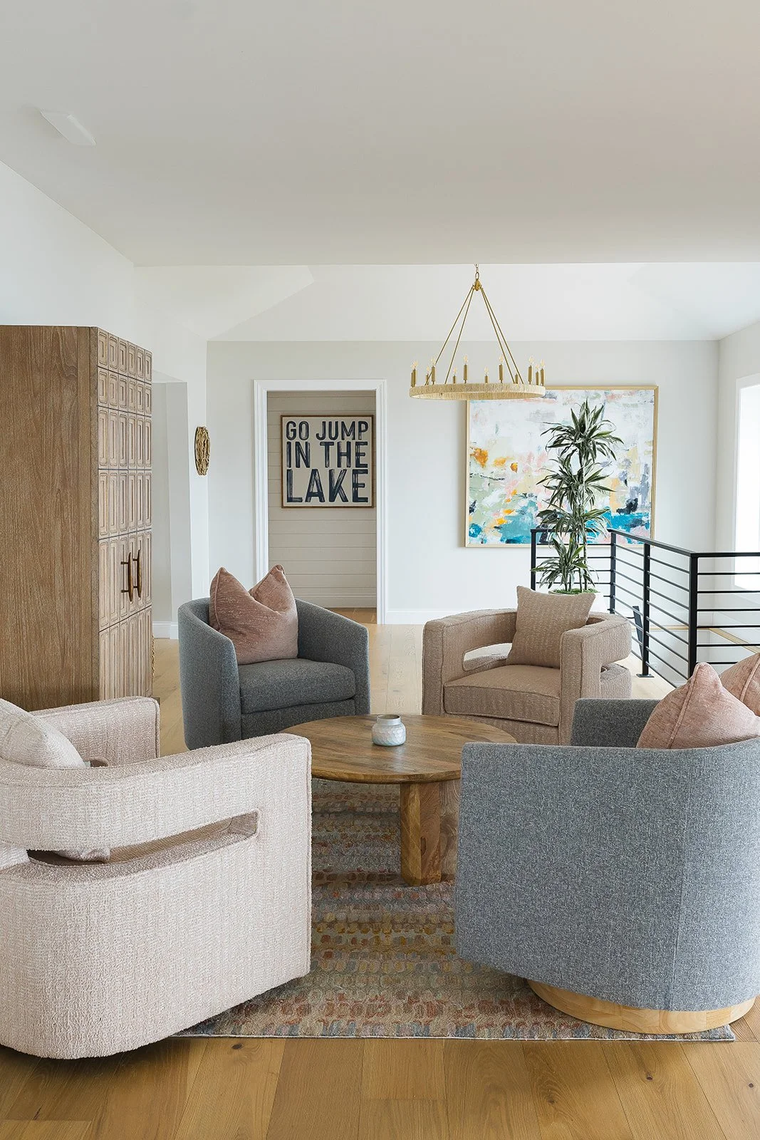 Living room with four armchairs in neutral and muted tones around a round wooden coffee table, a large wooden cabinet on the left, a framed sign on the back wall reading 'Go Jump in the Lake,' a colorful abstract painting, and a potted plant near a r