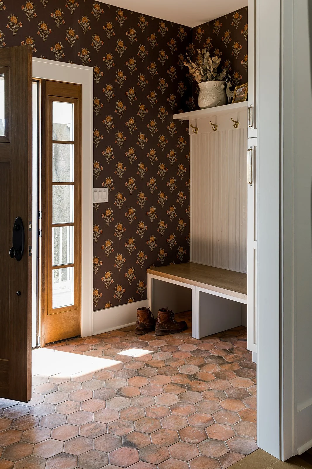 Entryway with hexagonal terracotta floor tiles, a wooden front door with glass panes, floral wallpaper on the wall, and a white built-in bench with hooks on the wall and a decorative vase on the shelf.