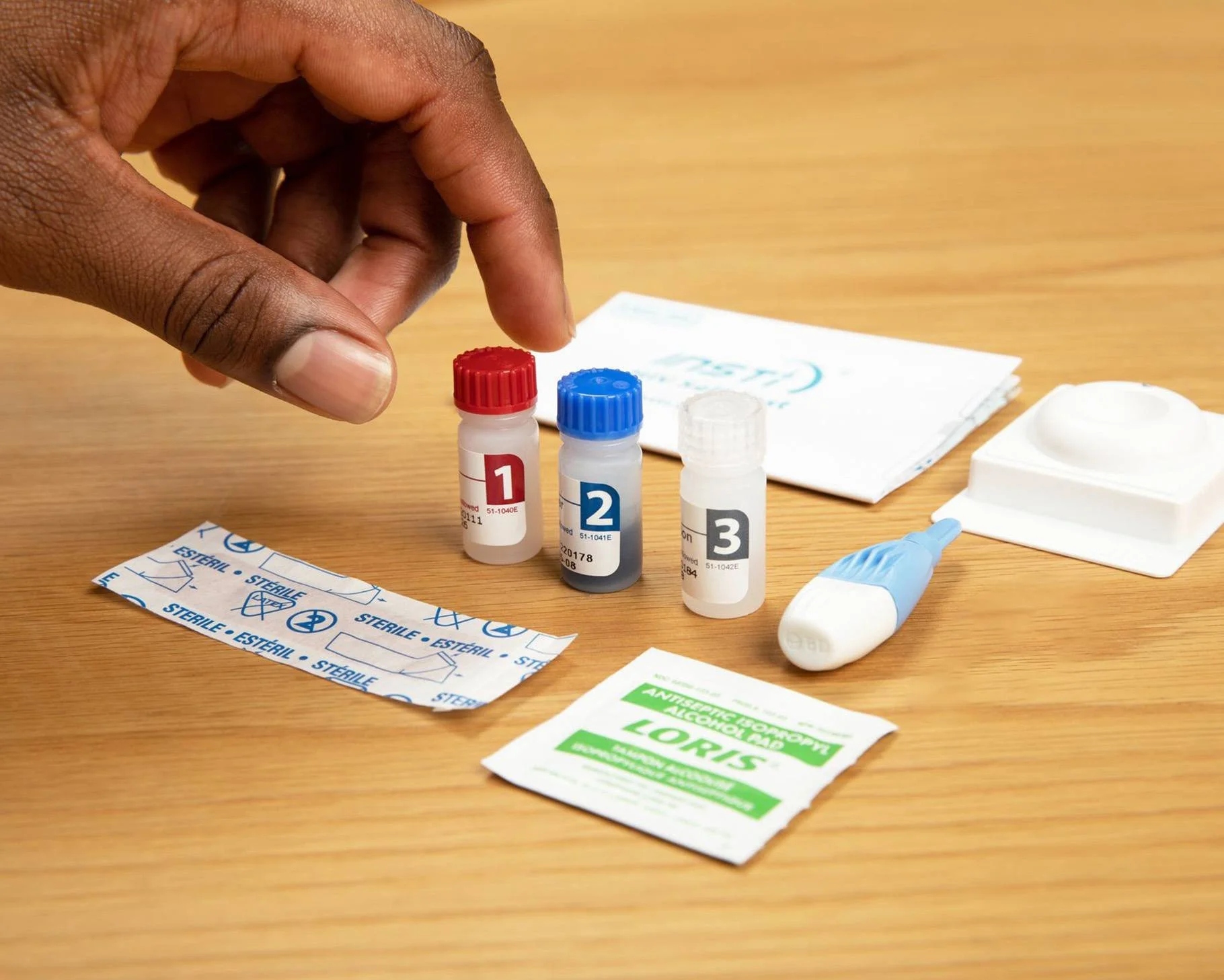 Hand placing a test tube labeled '1' on a wooden table, surrounded by medical supplies including vials, a thermometer, alcohol pad, and a packet of Loris antiseptic.