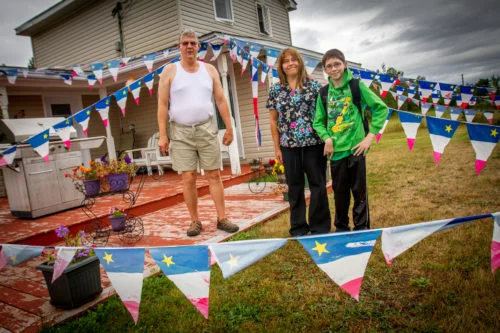 J.C. Benoit poses in front if his home in Tabusintac, NB, alongside his caregiver Chantal Breau and her son Jonathon Breau who decorated Benoit's home for the celebrations of National Acadian Day.