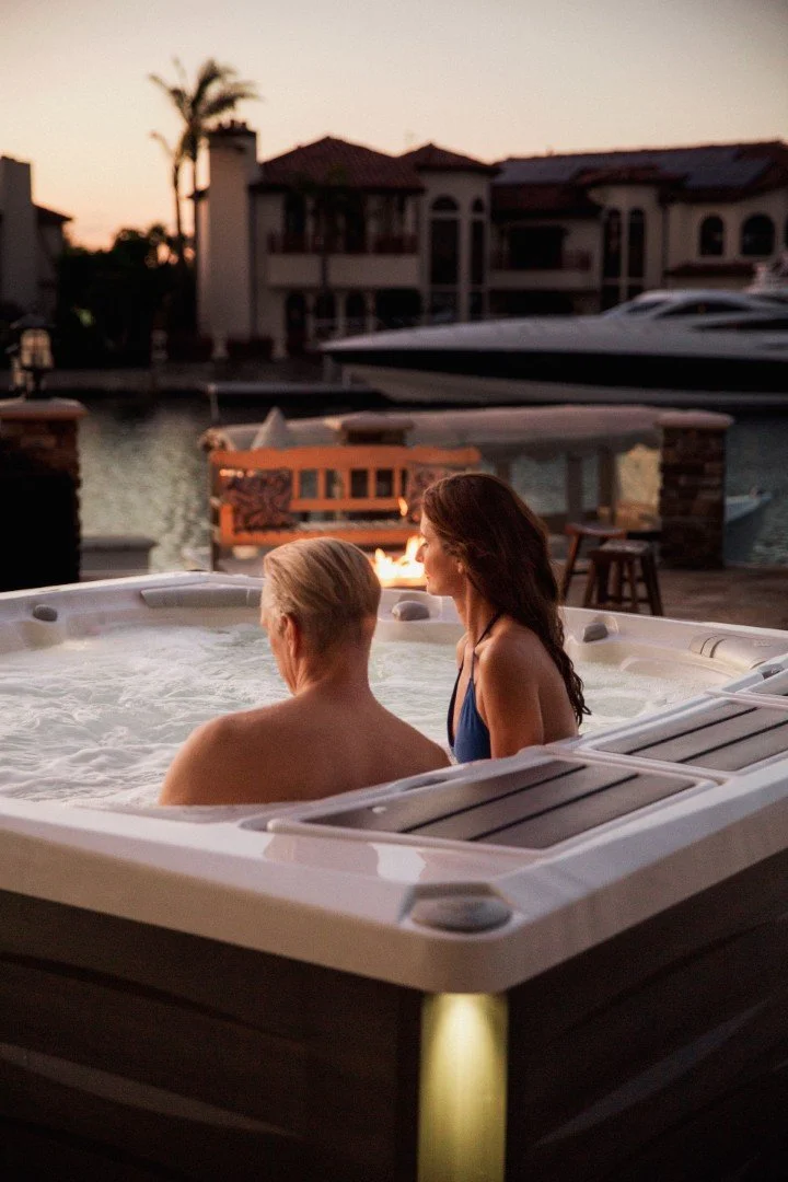 Two women relax in a hot tub on a dock during sunset, with luxurious houses and boats visible in the background.