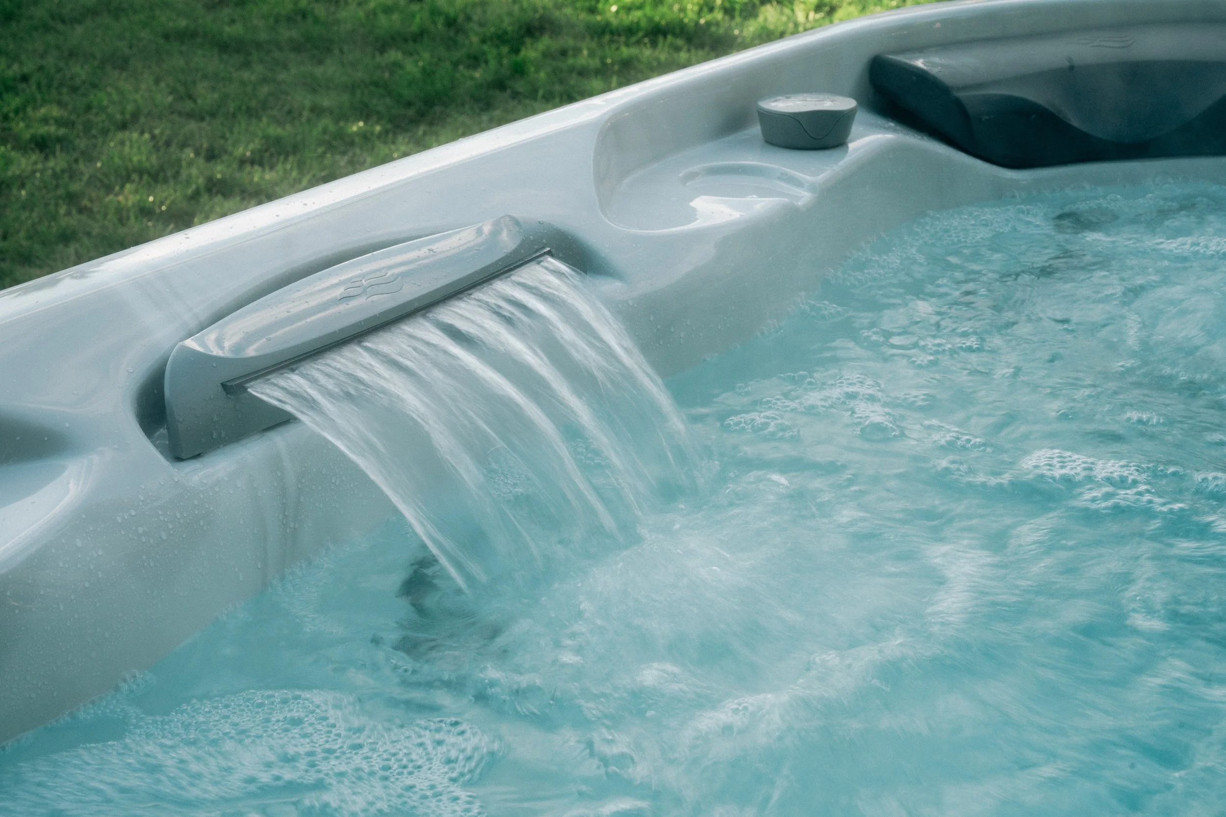 A hot tub with water flowing from a spillover feature into the tub, located outdoors on a grassy area.