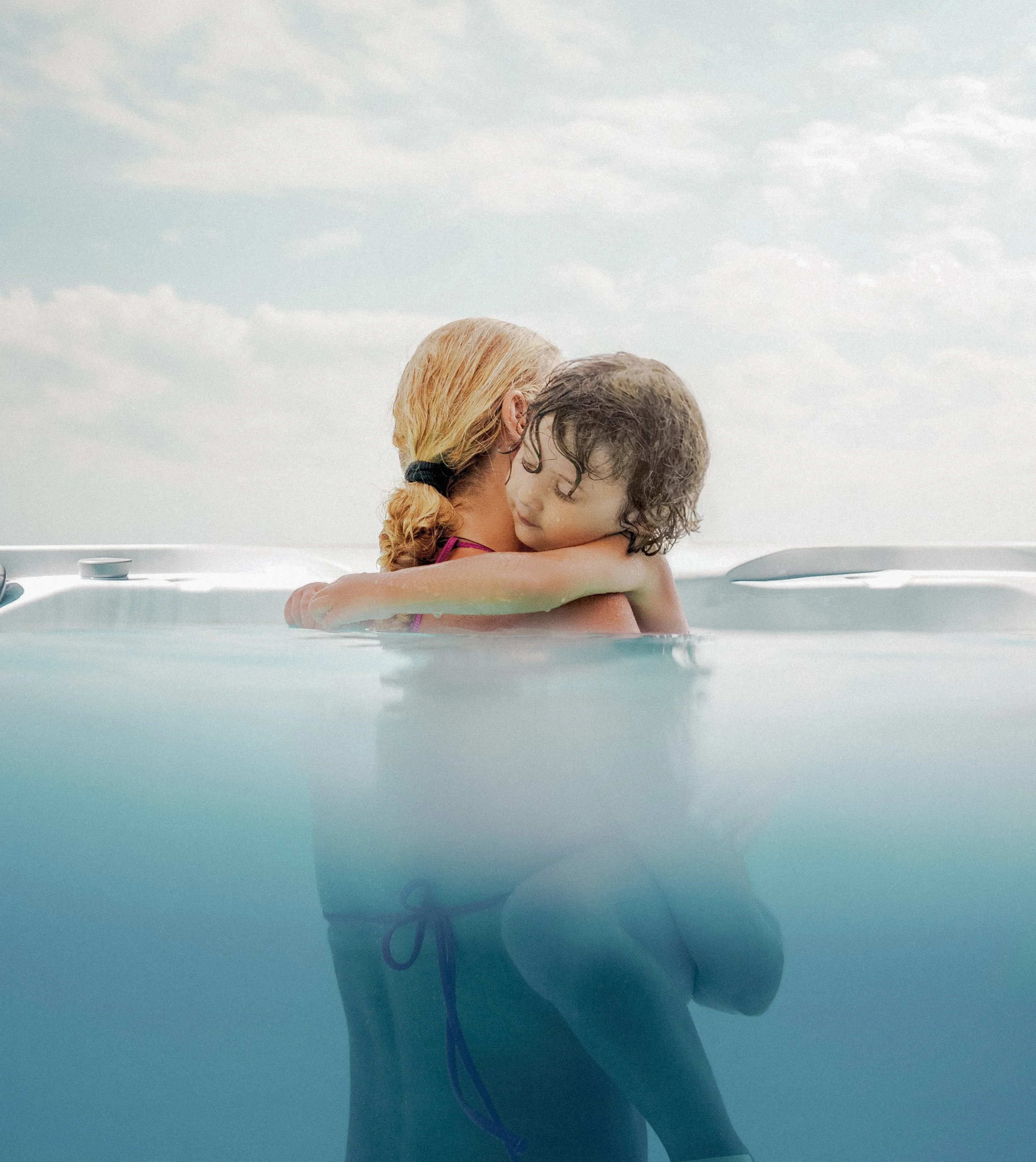 Two children, a girl and a boy, hugging in a hot tub outdoors with cloudy sky in the background.