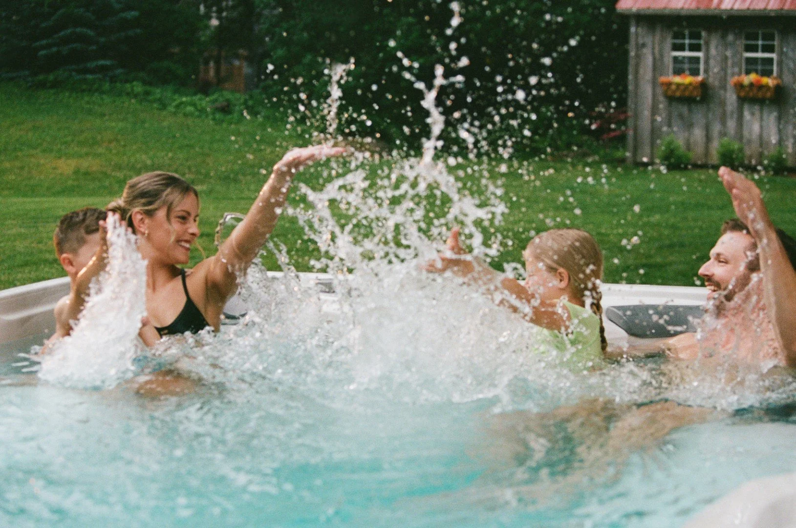 A family playing and splashing in a backyard swimming pool with a grassy lawn and a wooden shed in the background.