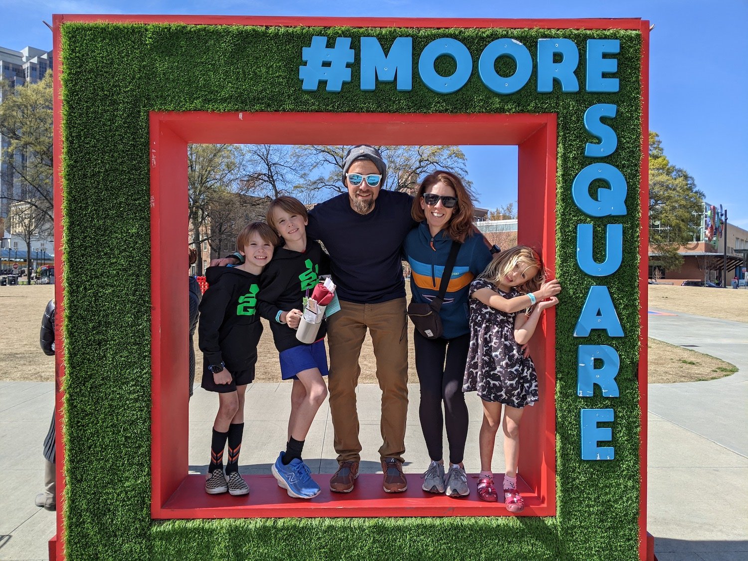 A family of four posing in a square outdoor frame with artificial grass and blue letters spelling “#MOORE SQUARE.” The background shows a park with trees and buildings.