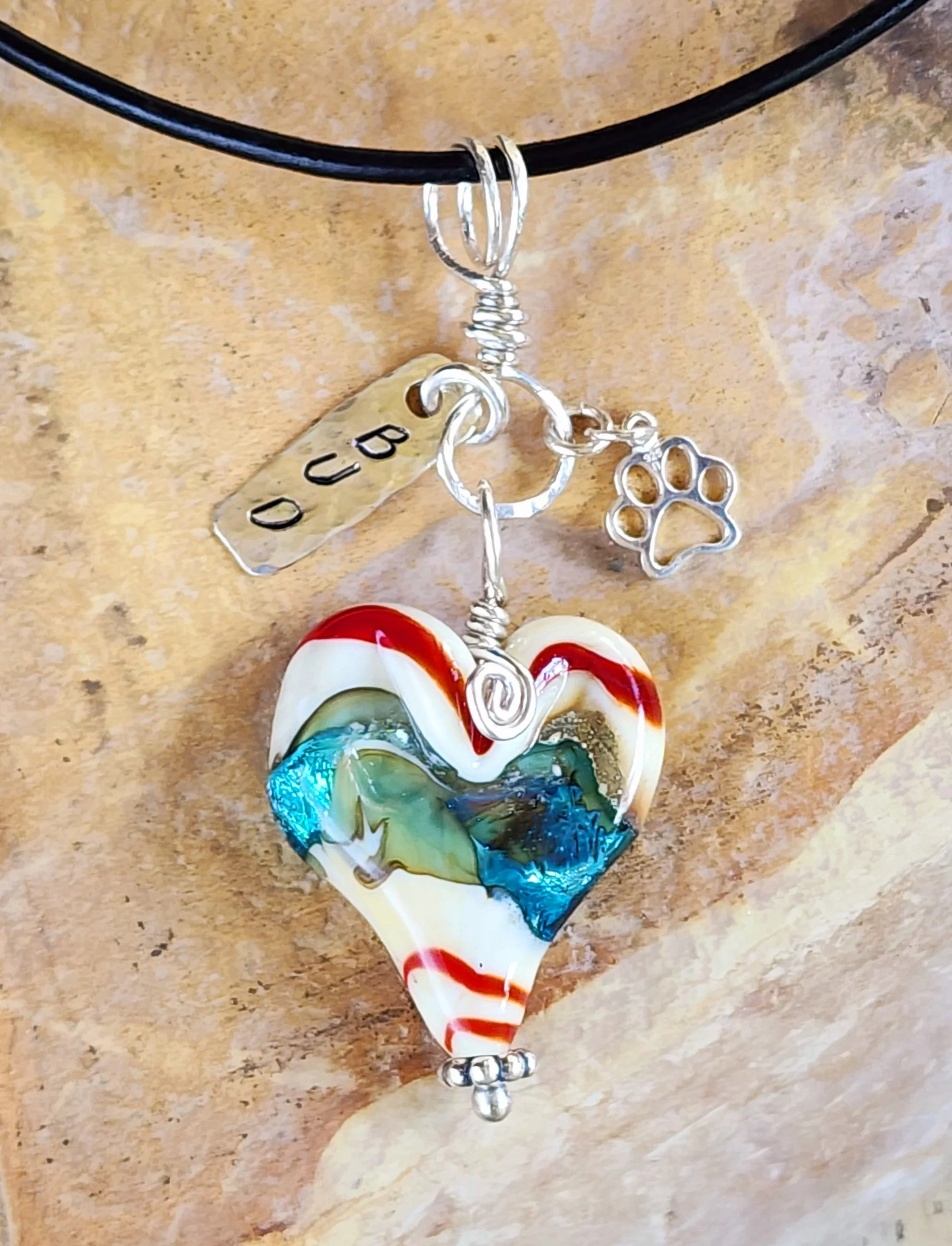 Handcrafted glass heart memorial pendant with red and blue swirls, featuring a stamped 'BUD' name tag and silver paw print charm.