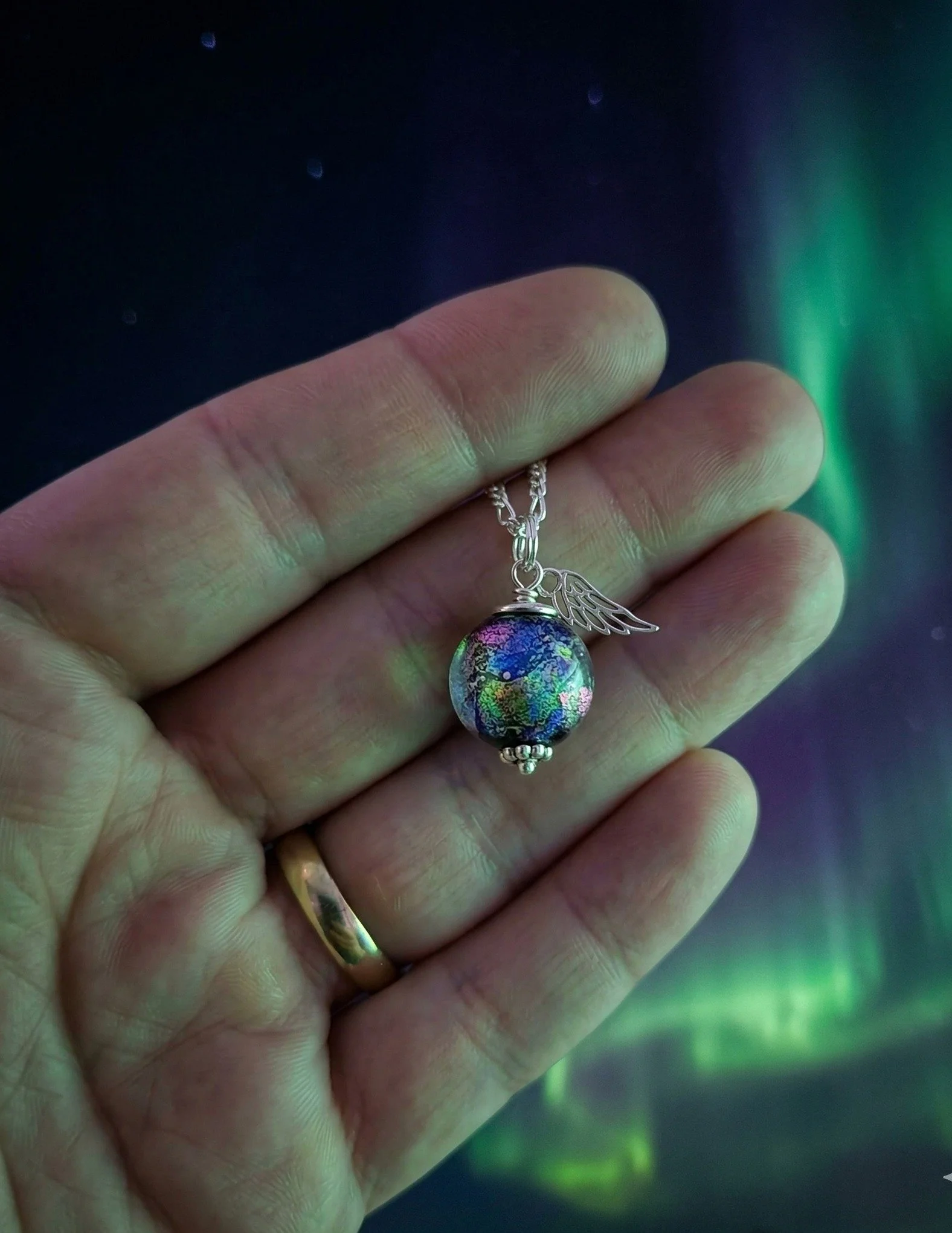 Hand-crafted petite memorial necklace featuring a glass galaxy bead and silver angel wing, held against a vibrant Montana Northern Lights night sky
