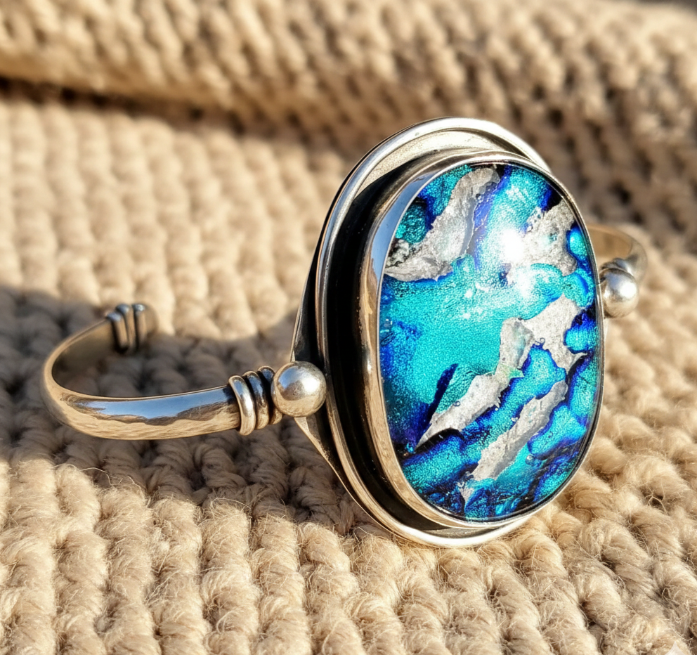 Large handcrafted sterling silver memorial cuff bracelet with a vibrant blue and silver iridescent glass stone, resting on a soft, chunky oatmeal-colored knit background in warm sunlight