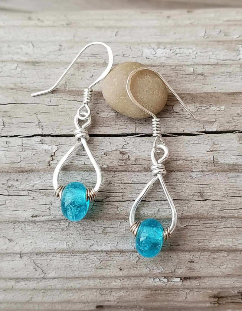 Handcrafted sterling silver teardrop memorial earrings with bright teal Sacred Trace glass beads, displayed on a natural light wood background as a delicate tribute for a cherished person.