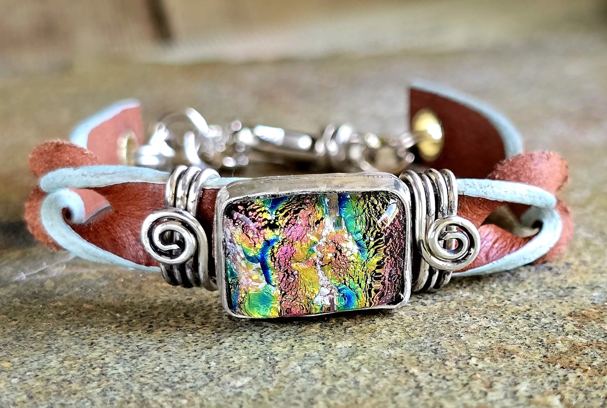 Close-up front view of an artisan memorial bracelet with a bezel-set dichroic glass stone, detailed silver wire work, and leather bands, displayed on a textured natural stone surface.