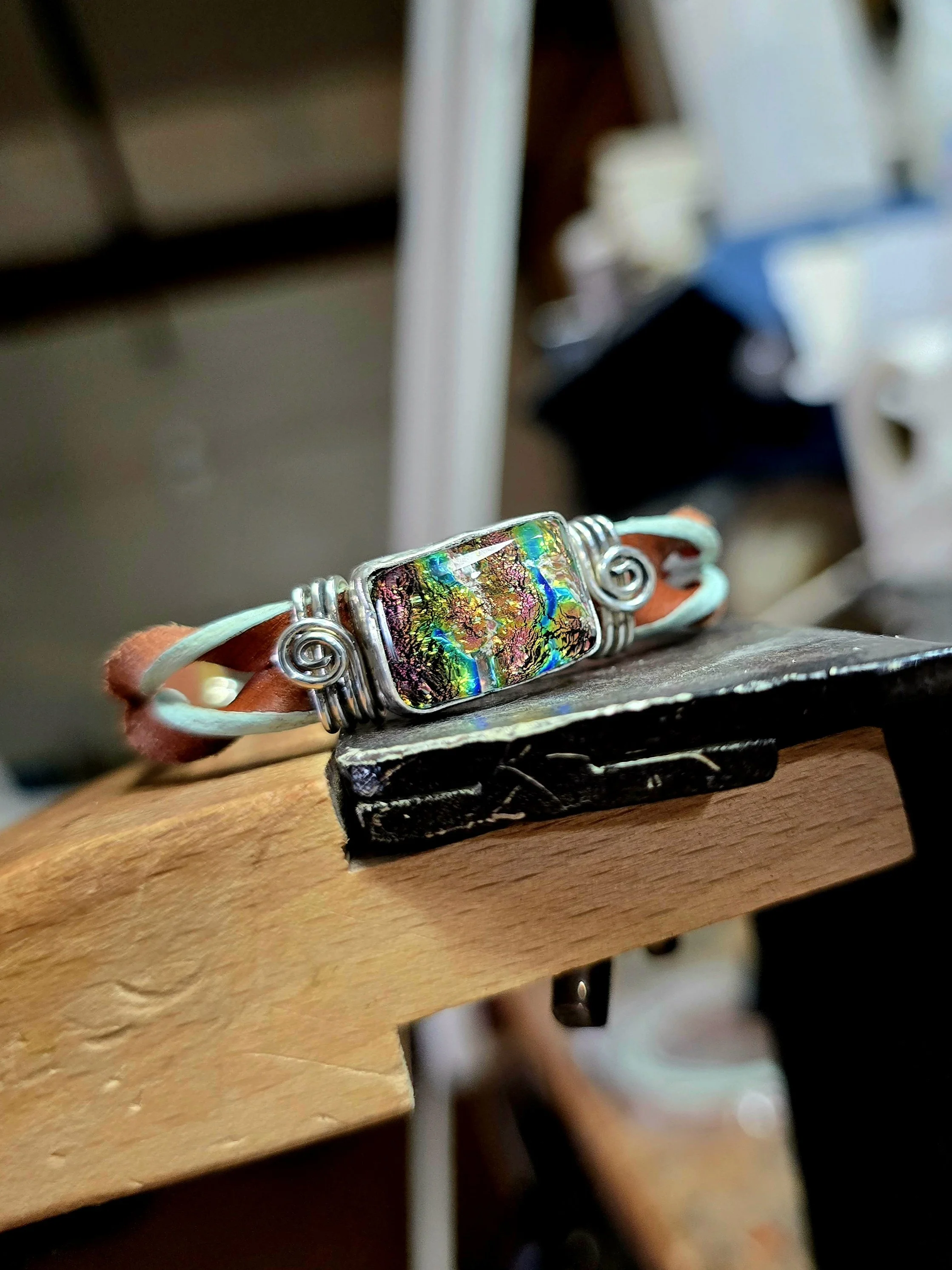 Handmade memorial bracelet featuring a rectangular, iridescent dichroic glass stone in a silver wire setting on a leather strap, resting on a rustic wooden workbench.