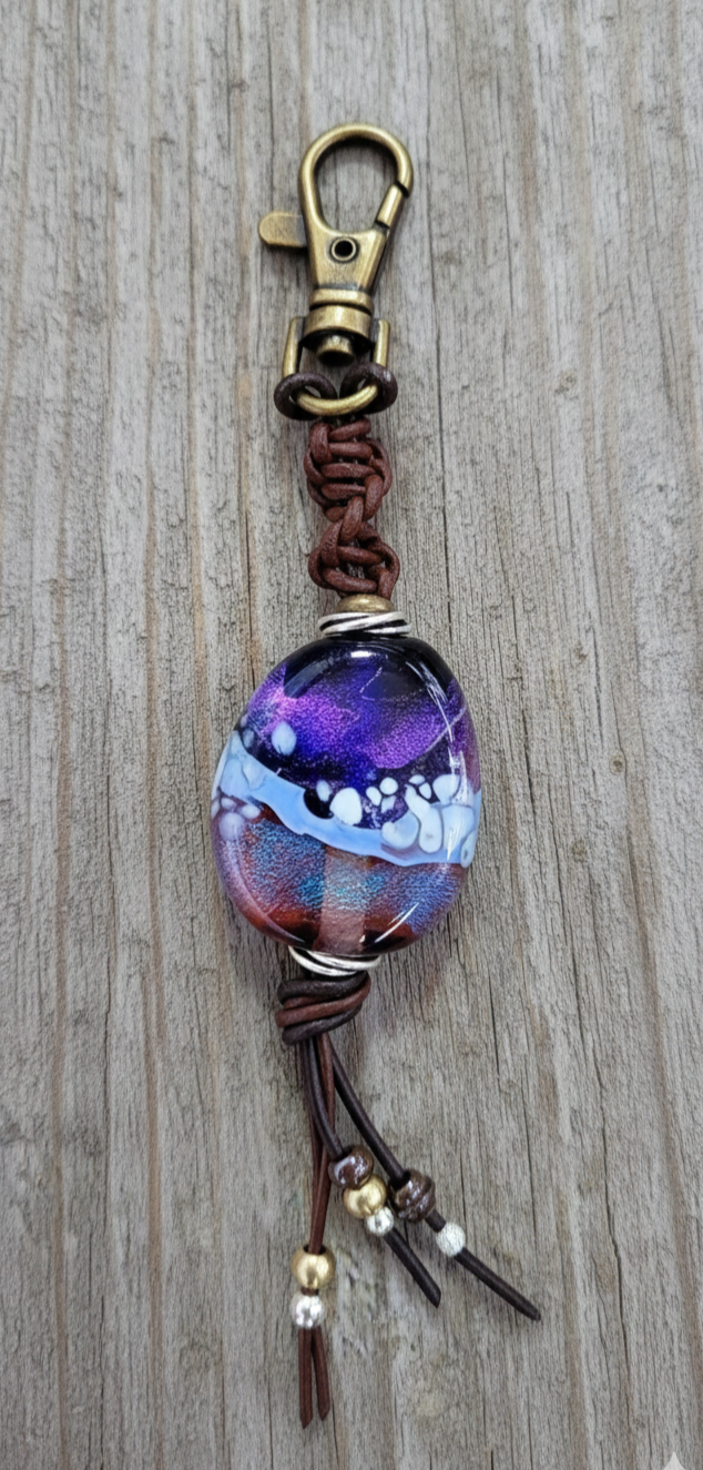 Handcrafted memorial glass token with a purple and blue Sacred Trace infusion, featuring a leather-braided clip, displayed on weathered gray barn wood as a comforting tribute for a cherished loved one.