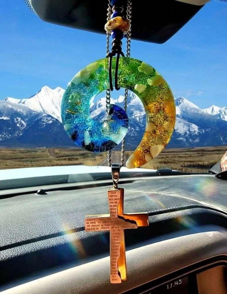 Hand-blown glass infinity spiral memorial car charm with colorful swirls and a leather hanging cord, shown against a blurred scenic Montana mountain landscape