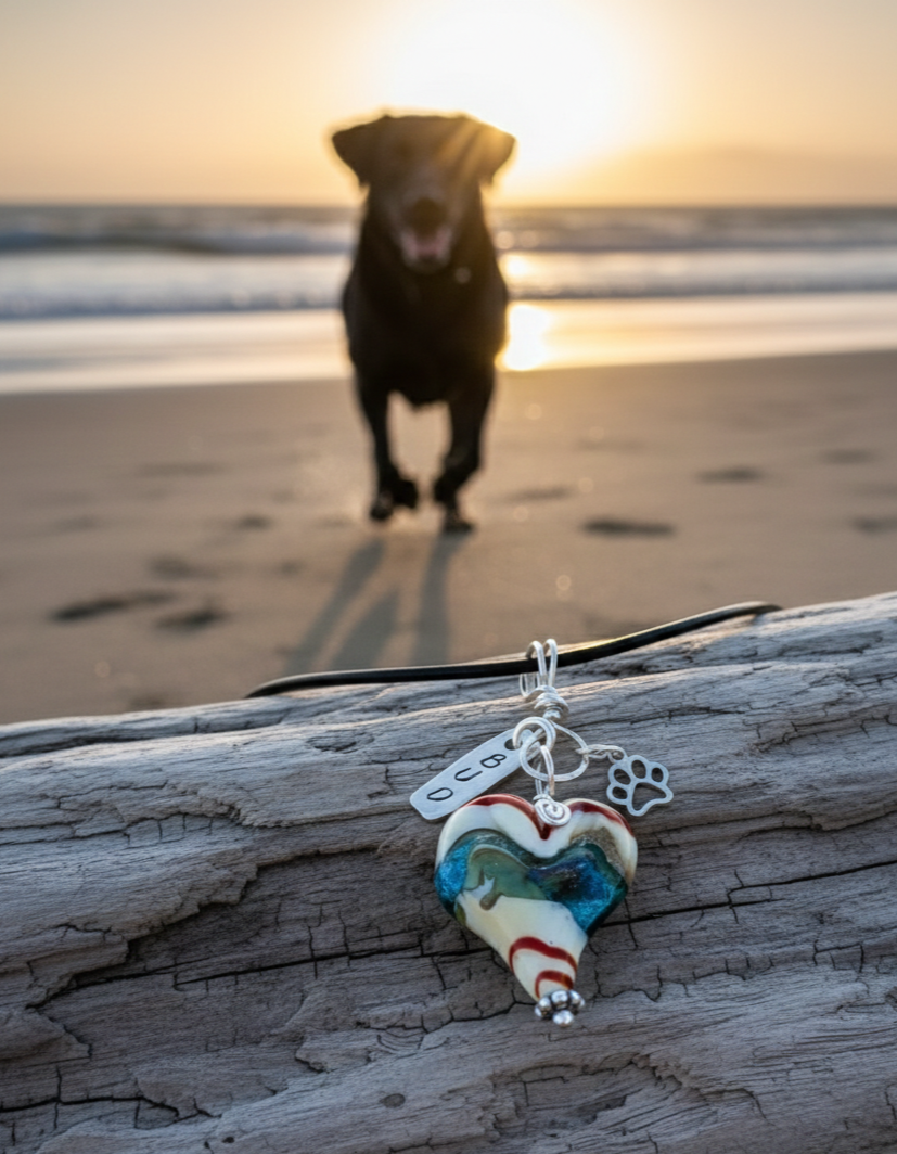 A glass heart pet memorial pendant with a 'Bud' tag draped over driftwood, with a black Lab running on a sunlit beach in the soft-focus background