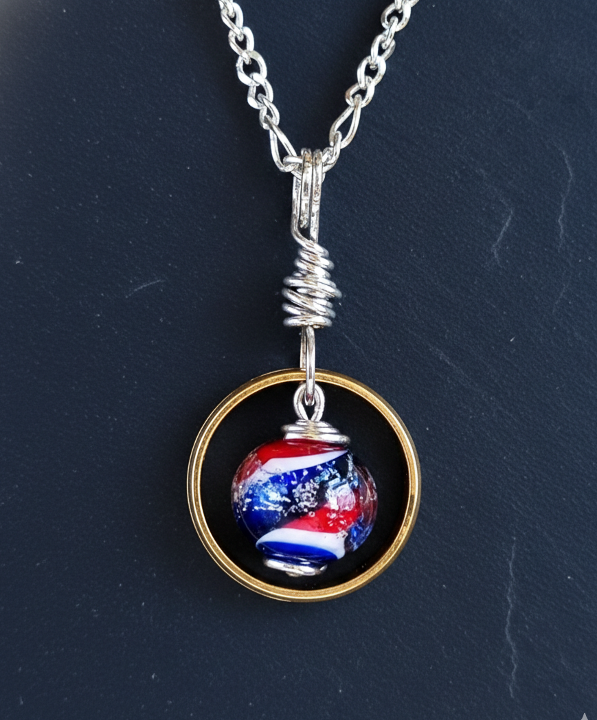 A handcrafted memorial necklace featuring a vibrant red, white, and blue swirled glass bead suspended inside a gold wedding ring, displayed on a dark blue slate background.