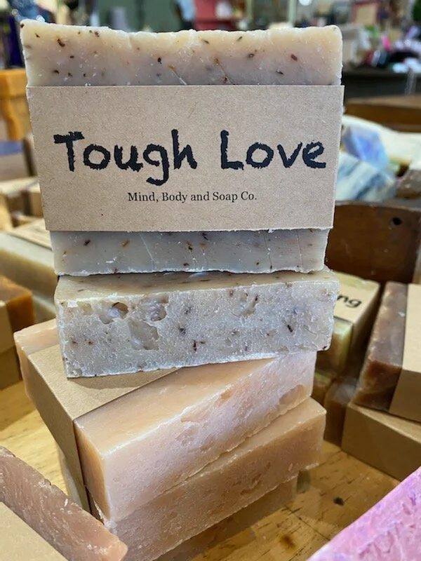 A photo of a rectangular block of soap with a tan label wrapped around it. The soap is a creamy color with a smooth surface and has a simple design.
