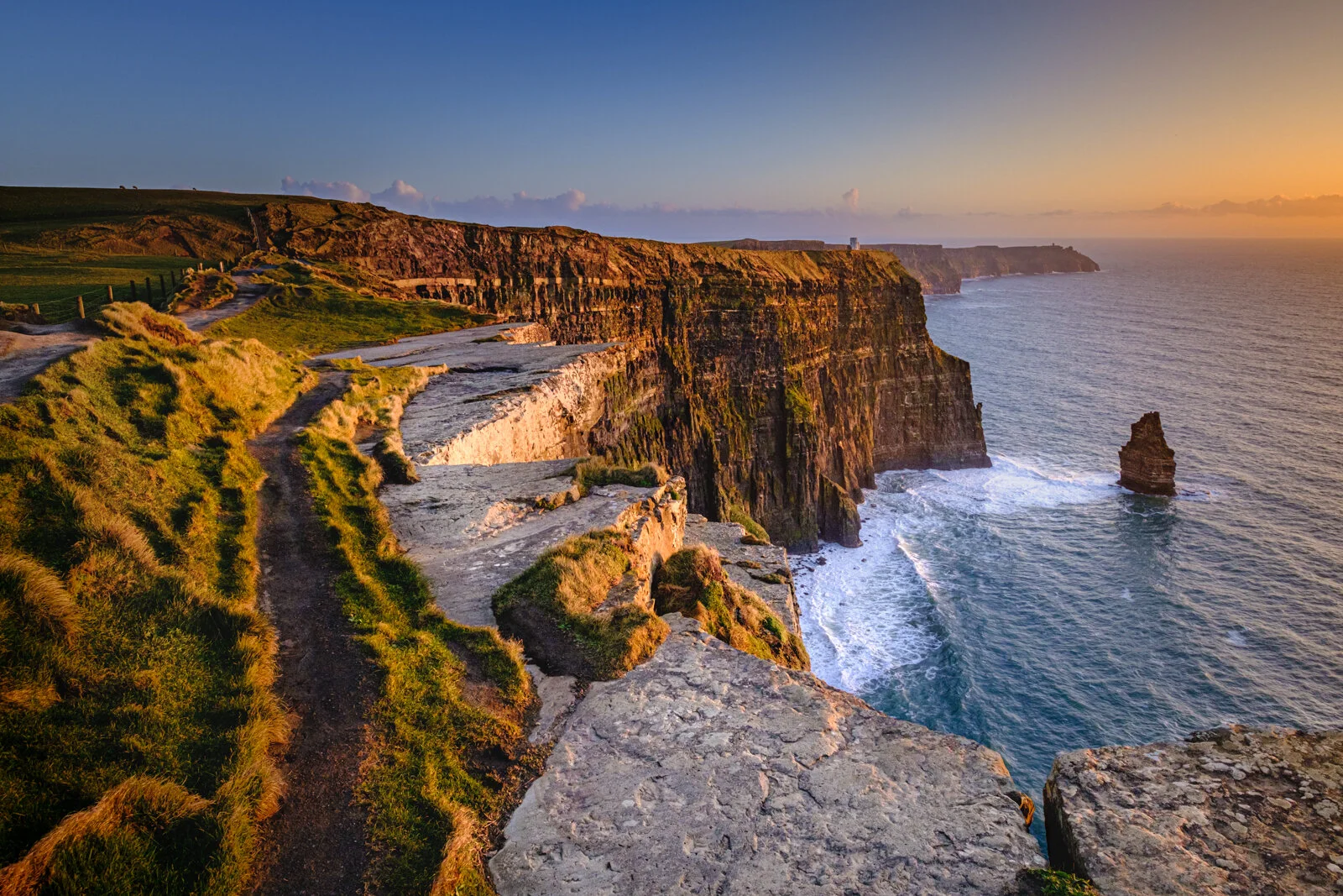 Cliffside trail along the Irish coast at the Cliffs of Moher at sunset, with rugged cliffs and ocean waves below.