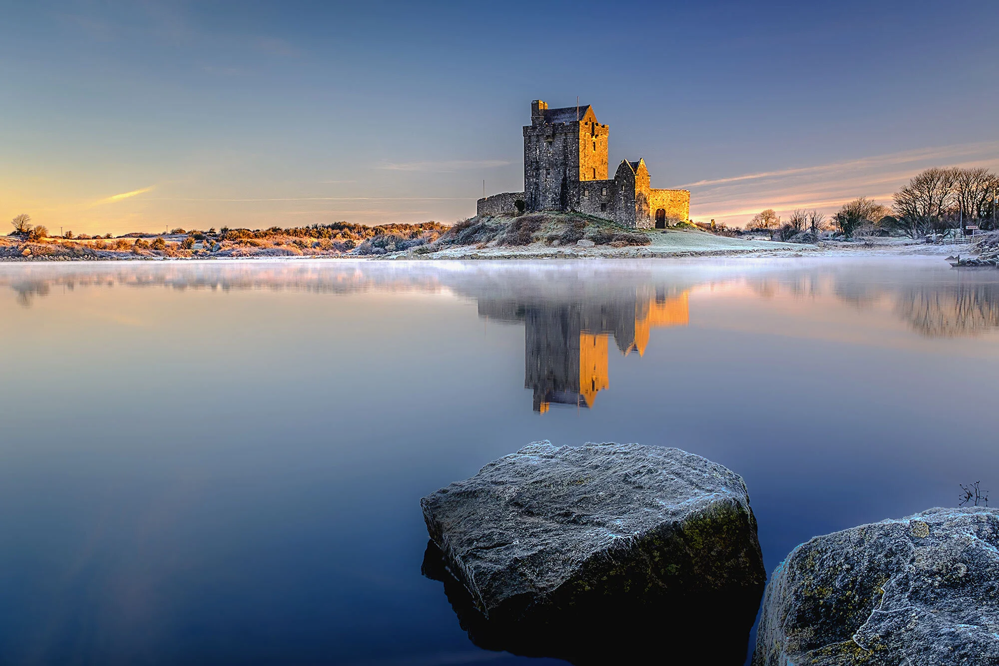 A castle on a small island reflected in a calm, partially frozen lake during sunrise or sunset with rocks in the foreground.