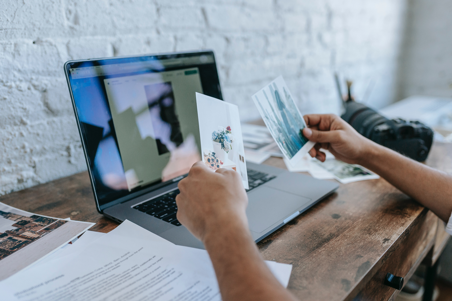 Person holding printed photographs while working on a laptop at a wooden desk with papers and a camera in the background.