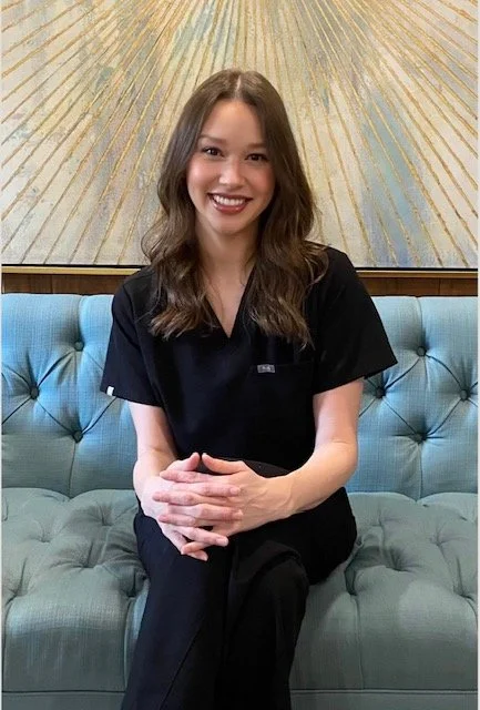 A wSoman sitting on a blue tufted couch, smiling, with tattoos on her arms. She is wearing a black uniform with a name tag and a logo on the shirt. Behind her is a decorative artwork with golden rays.
