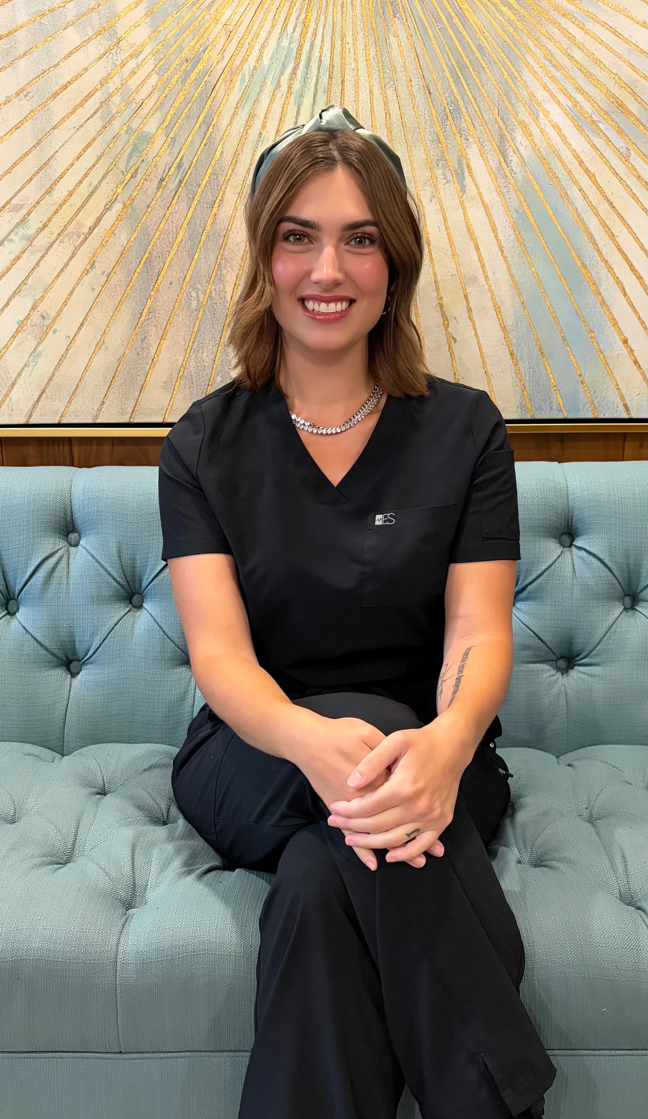 Person sitting on a blue tufted sofa in front of a patterned wall, wearing a black outfit and a name tag.