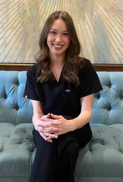 A wSoman sitting on a blue tufted couch, smiling, with tattoos on her arms. She is wearing a black uniform with a name tag and a logo on the shirt. Behind her is a decorative artwork with golden rays.