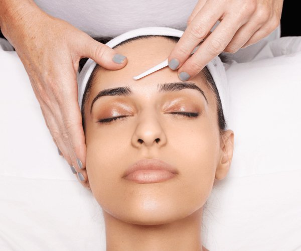 Gloved hands applying a small blade to a woman's forehead as she lays with her eyes closed
