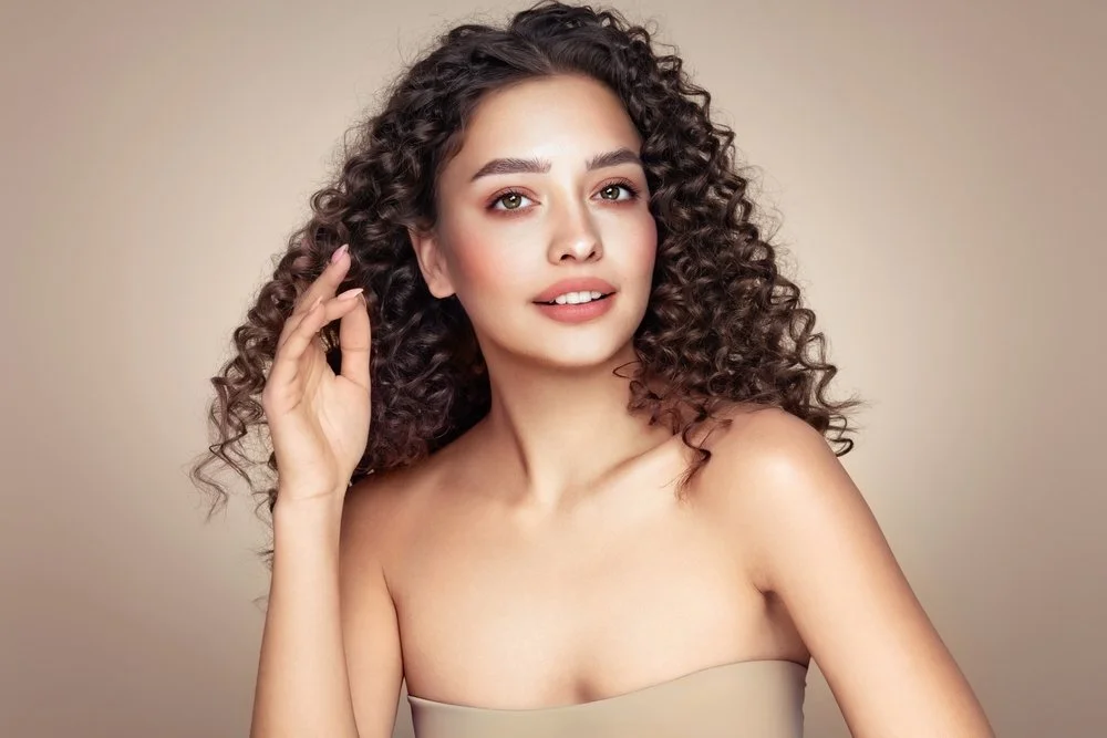 A light-skinned woman with dark, curly hair smiling at the camera as she brushes her hair away from her face