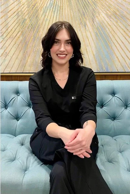 Person sitting on a blue tufted sofa in front of a patterned wall, wearing a black outfit and a name tag.