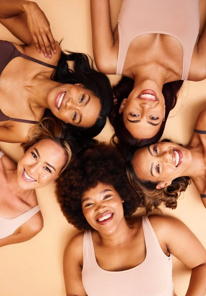 Five women of different ethnicities laying on their ground with their heads together in a circle, smiling at the camera