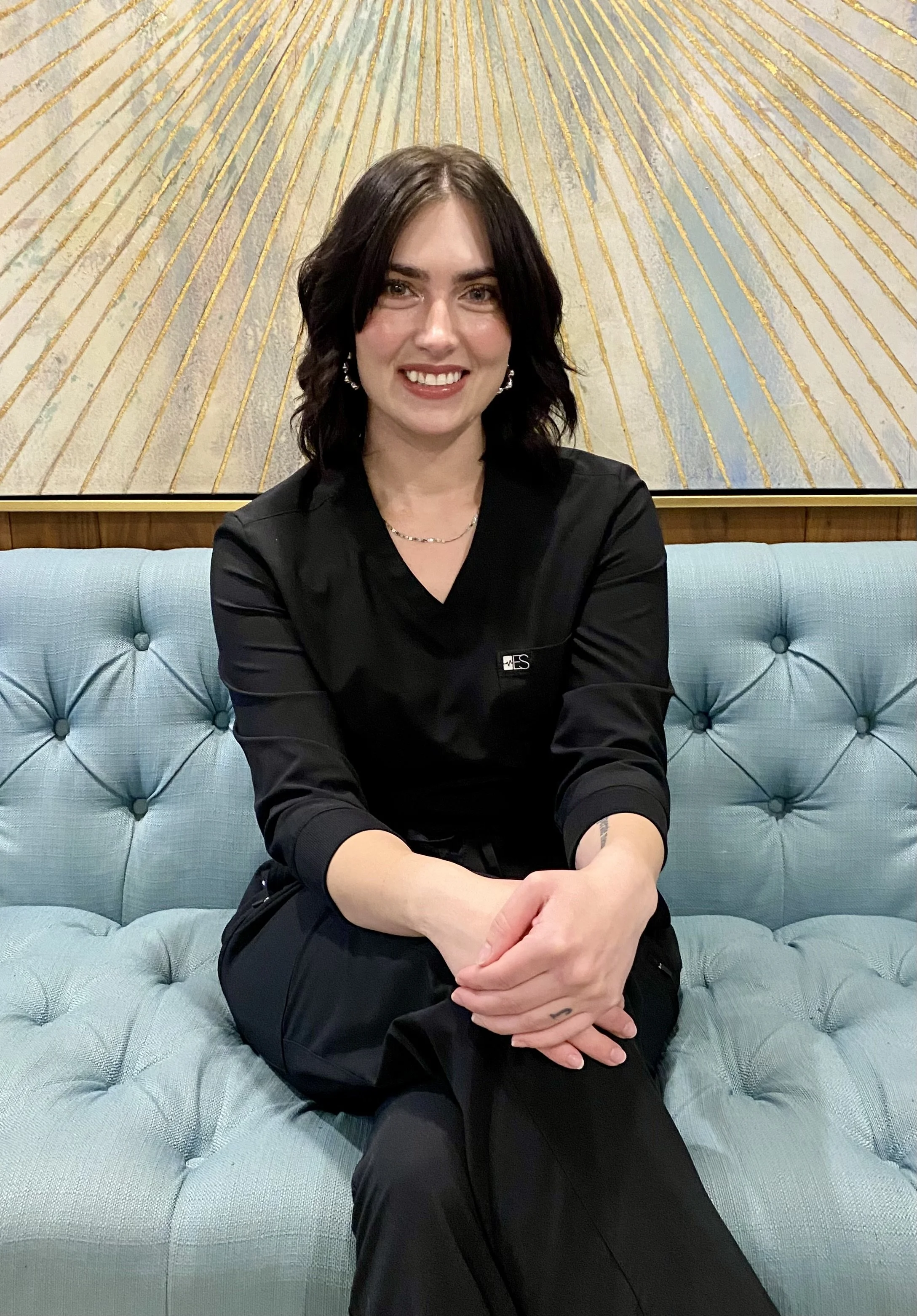 Person sitting on a blue tufted sofa in front of a patterned wall, wearing a black outfit and a name tag.