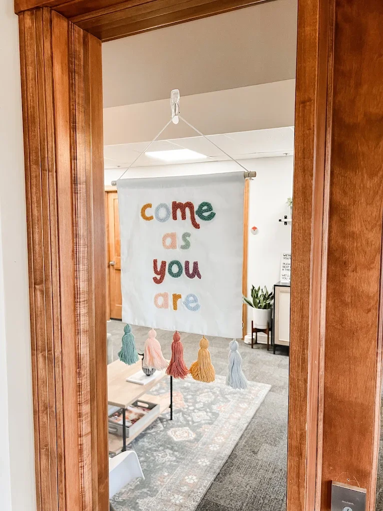 A decorative fabric banner hanging on a door frame with colorful embroidered letters spelling 'come as you are', decorated with pastel-colored tassels hanging from the bottom.