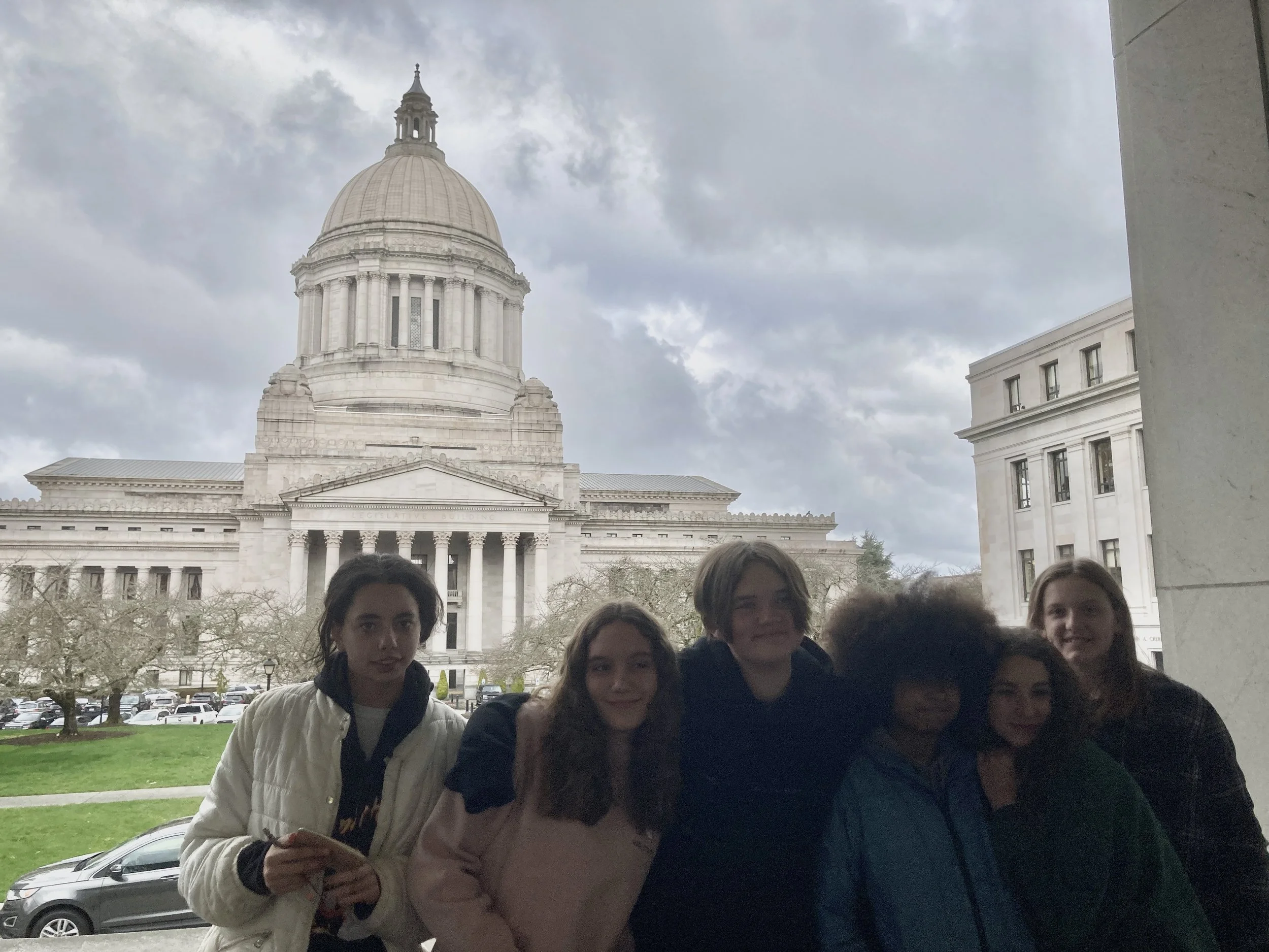 Mountain landscape with snow-capped peaks in the background, rolling grassy hills in the foreground, and a modern wooden building nestled on the hillside.