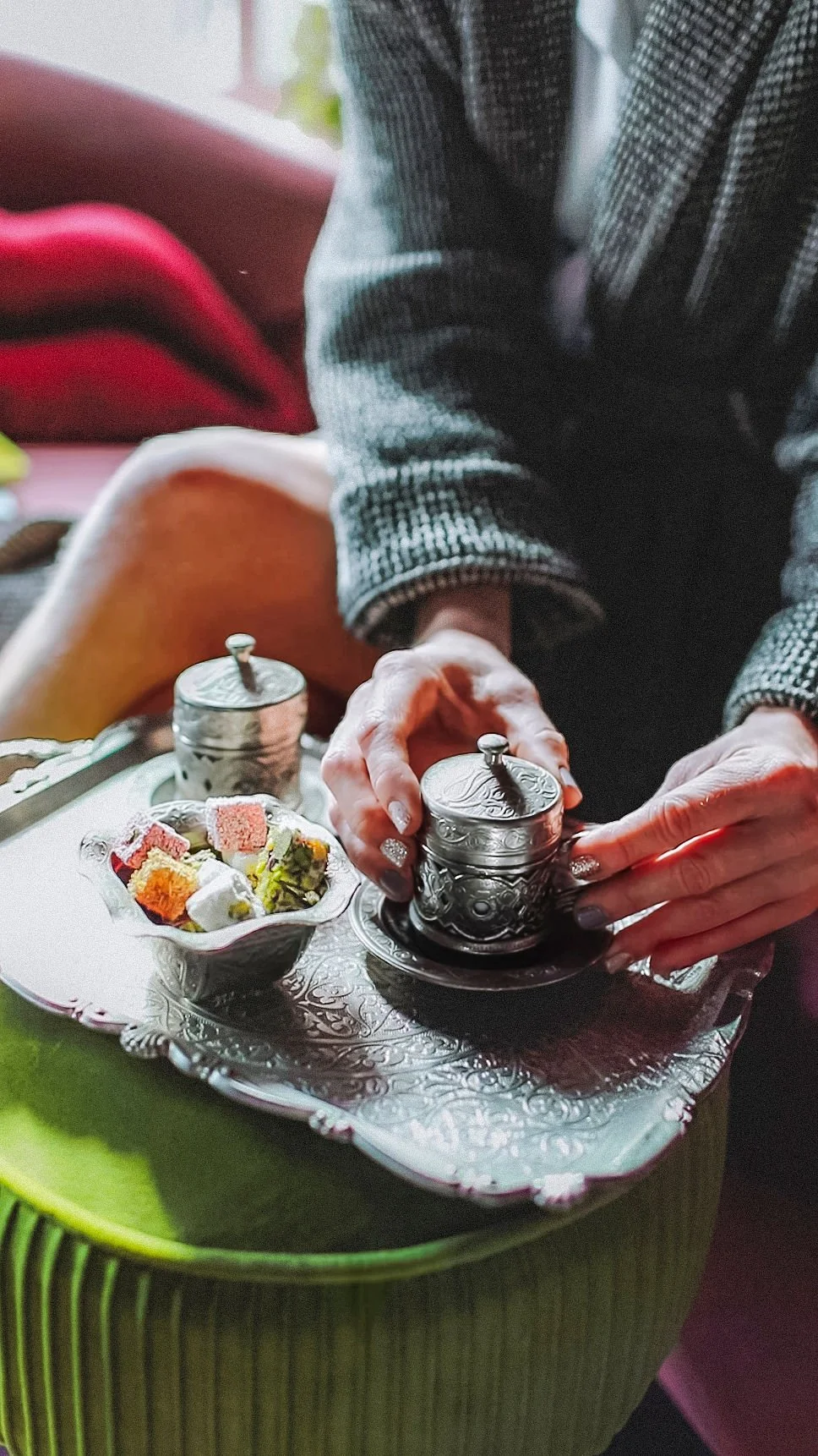 Person's hands holding a silver container with intricate designs, part of a tea set on a decorative tray with small colorful sweets, sitting on a green table wearing a wool house coat from Together Textiles.