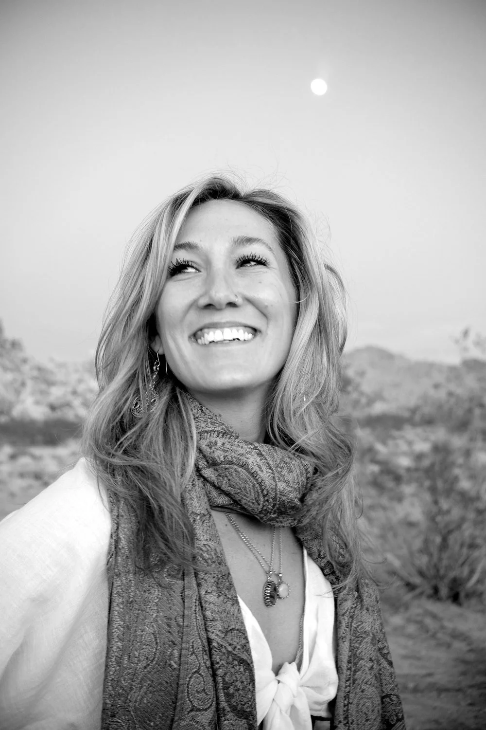 Black and white portrait of astrologer Sarah Paasch smiling up at the moon in the desert