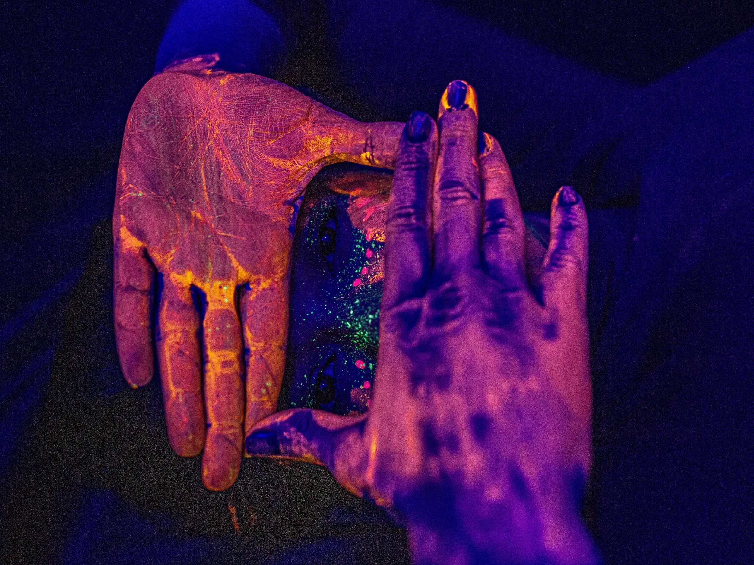 Hands covered in glowing neon paint cupping glitter under ultraviolet light