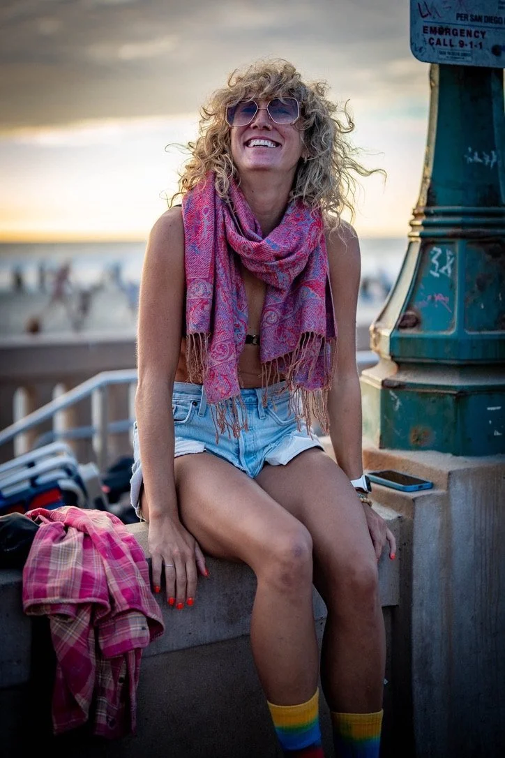 Astrologer Sarah Paasch smiling in a pink scarf, sitting on a beach boardwalk at sunset