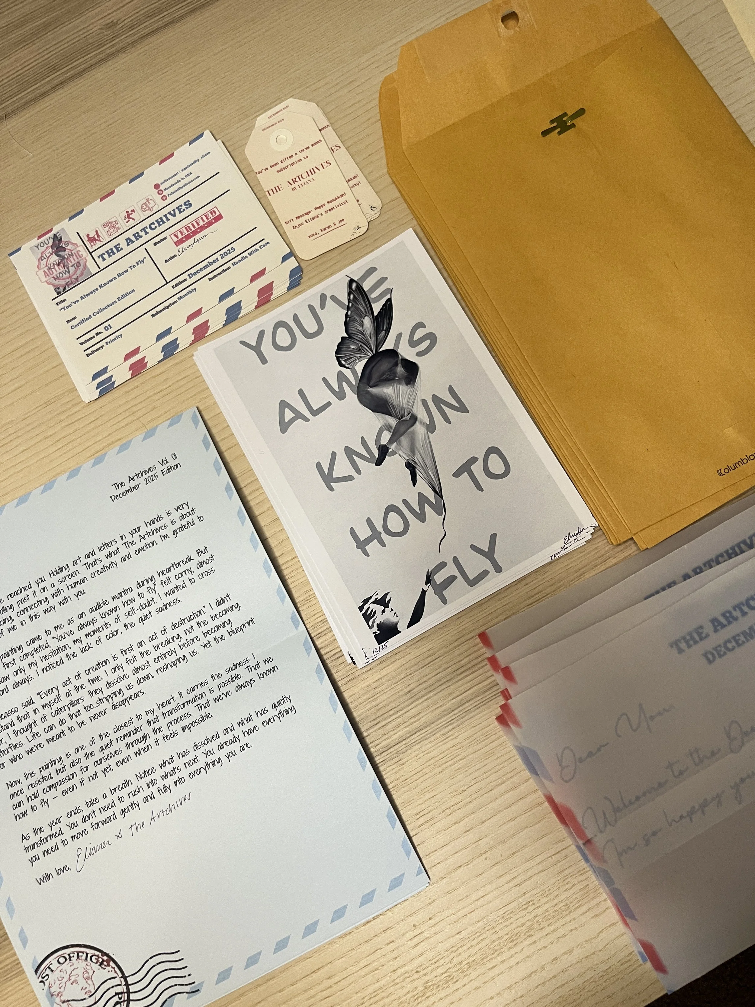 Collection of travel-themed stationery, including a letter, a set of vintage-style archives labels, a card with a butterfly illustration and motivational quote, and multiple manila envelopes on a wooden surface.