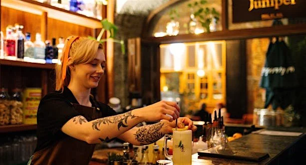 A woman with tattoos and short blonde hair preparing a drink behind a bar in a cozy, well-lit pub or restaurant.