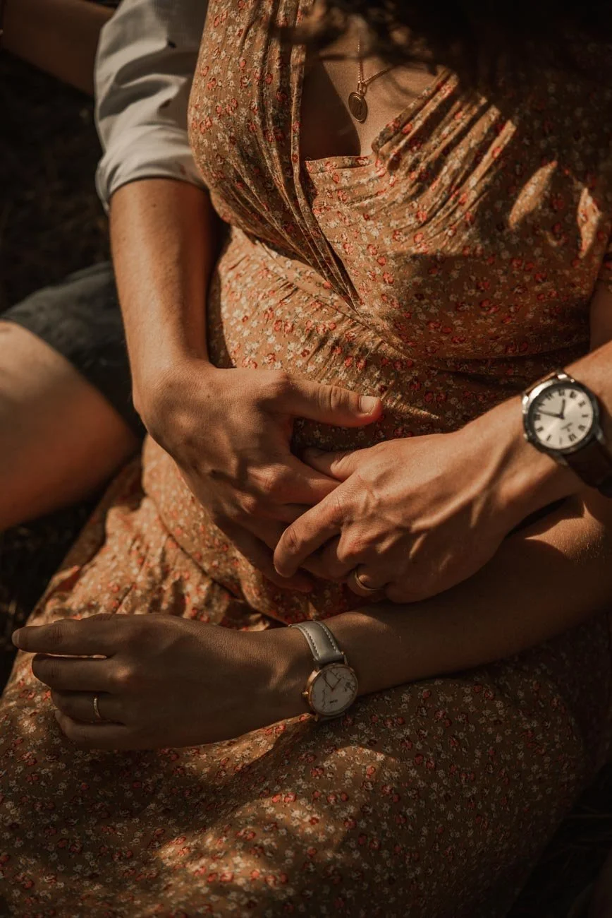 Deux personnes enlacées, une main posée sur le ventre, deux montres visibles, vêtues d'une robe à fleurs et une chemise blanche.