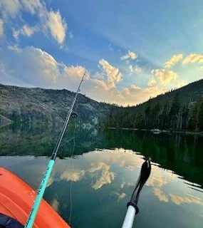Fishing rod extending into a calm lake with mountains and trees in the background, during sunset.
