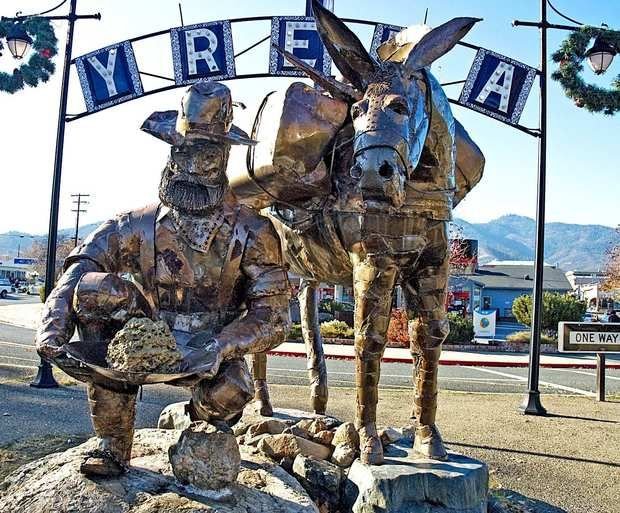 Large metal sculpture of two donkeys, one sitting and one standing, in front of an archway with a sign that reads 'YREKA'; a street with traffic and mountains in the background.