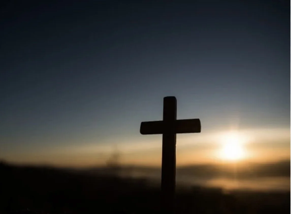 Silhouette of a cross against a sunset or sunrise sky over a body of water.
