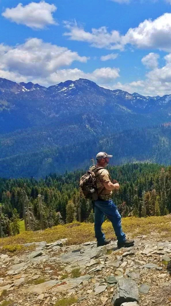 A man wearing camouflage and a backpack hiking on a rocky trail in a mountainous forest area with tall mountains and a partly cloudy sky in the background.