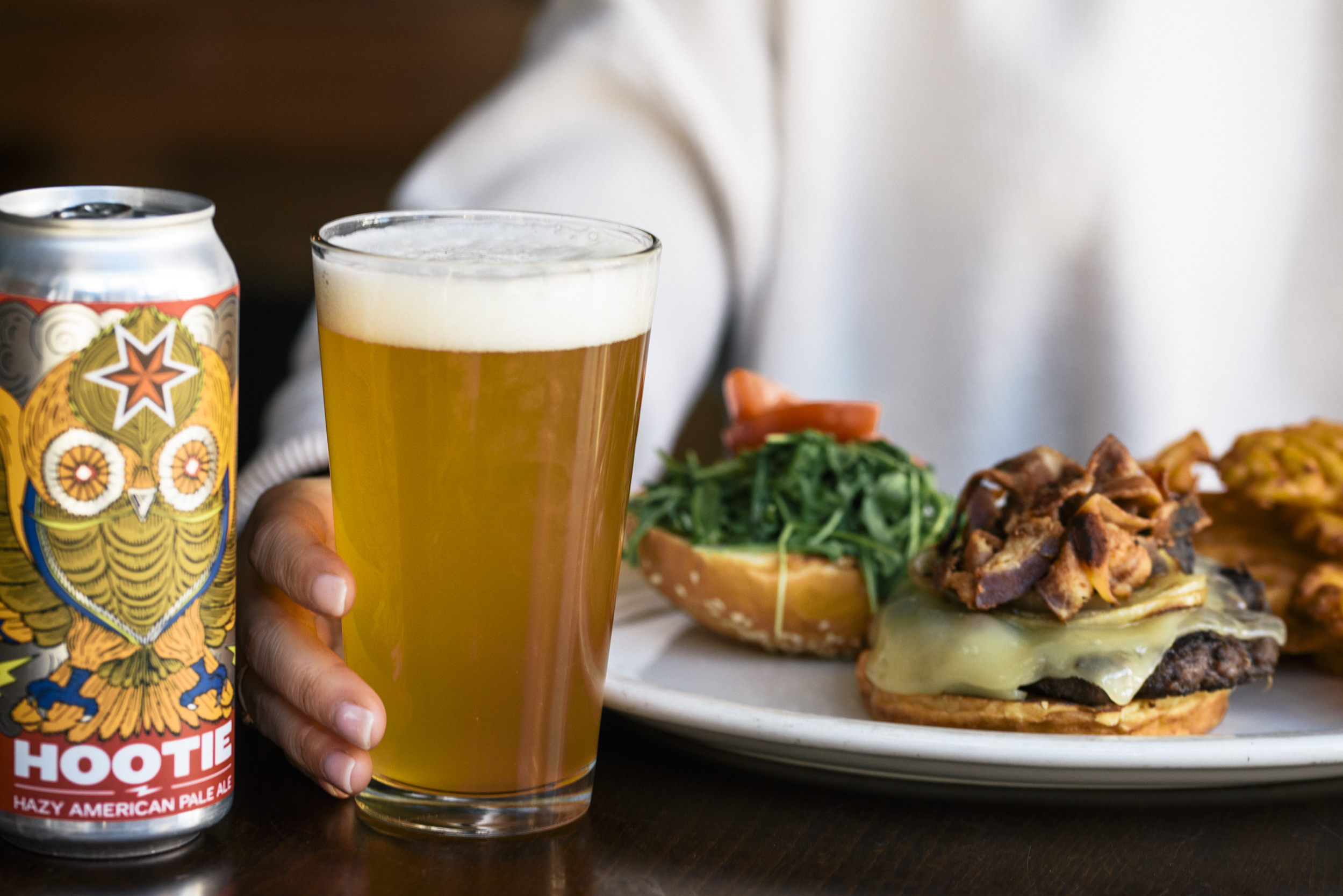 a close up of a woman with open bacon cheese burger on a plate infront of her with a pint of freshly poured Hootie beer next to plate and can of beer on left side of photo