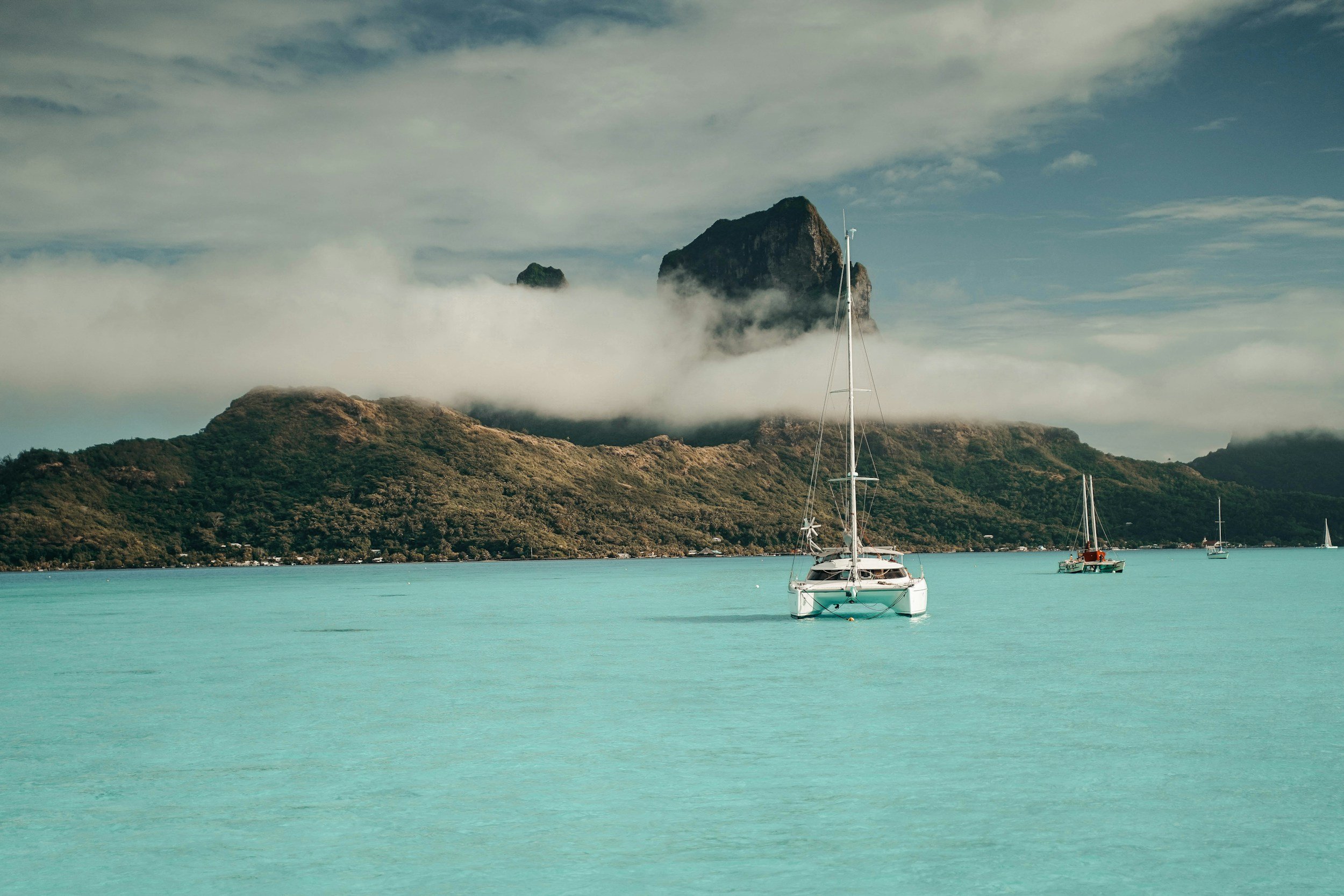Boats anchored in a turquoise bay with a lush green mountain and mist clouds in the background.